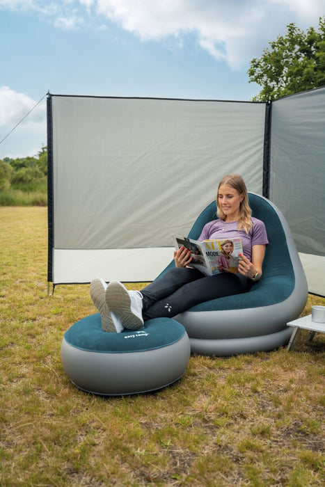 Easy Camp Comfy Lounge Set - Flocked Inflatable Camping Chair and footstool pouf Set lifestyle image of lady sat reading a magazine in chair with feet up on pouf, windscreen behind her and tress and blue sky with clouds in background. small coffee table half in image on right side with mug on it.