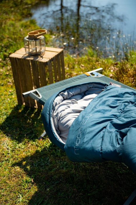Easy Camp Pampas  Folding Camp Bed - Pacific Blue lifestyle image of bed with moon 200 sleeping bag on by lake with wooden box with light on it