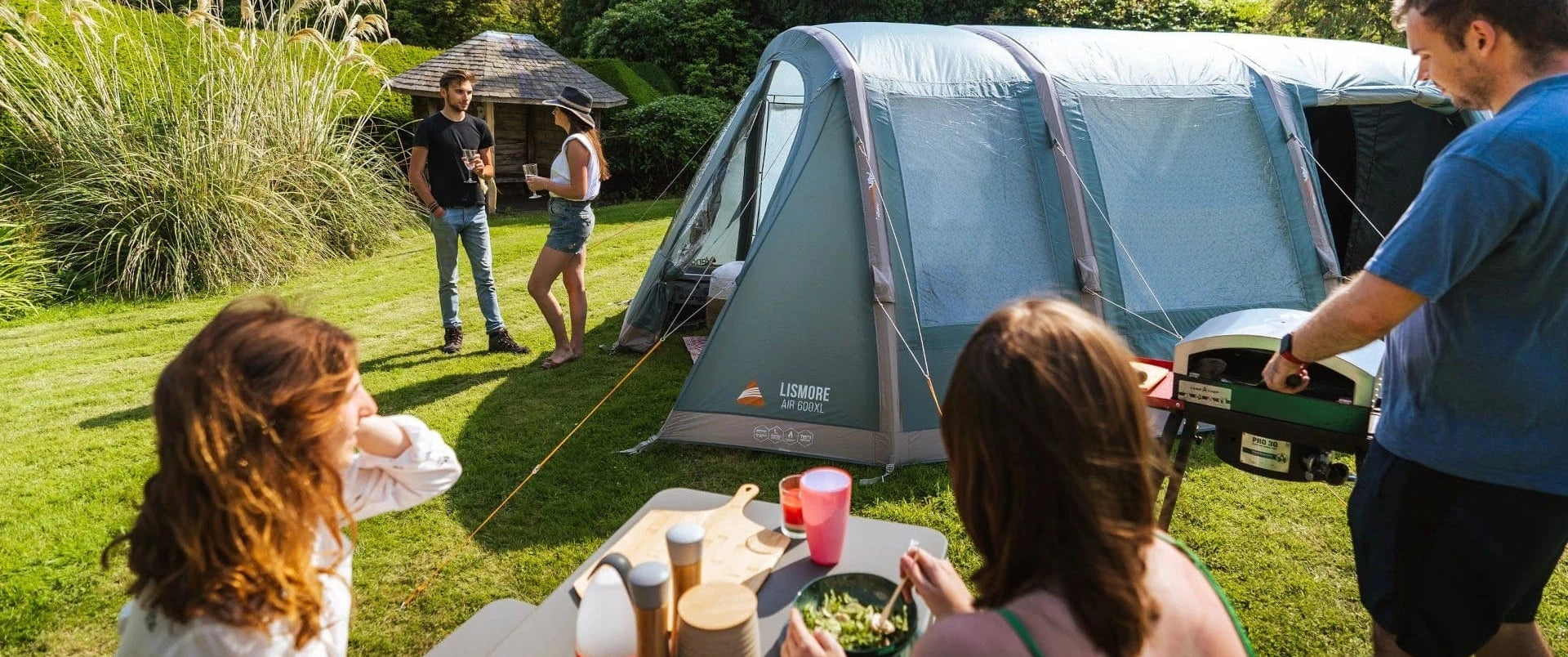 A family camping outdoors on a grassy lawn next to a large blue and gray Coleman tent, with a picnic table in the foreground and two people standing near the tent entrance.