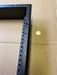 A close-up of a black metal mounting bracket or frame corner with a row of screw holes, shown on a brown cardboard surface next to a coin for scale.