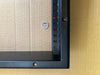 Close-up of a black metal picture frame corner with mounting holes, shown against a brown cardboard background with a coin for scale.