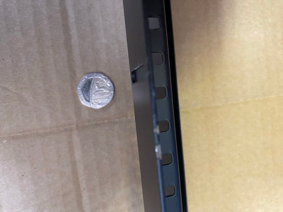 A close-up of a black metal mounting bracket or frame edge with rectangular cutouts, shown against a brown cardboard background with a small silver coin for scale.