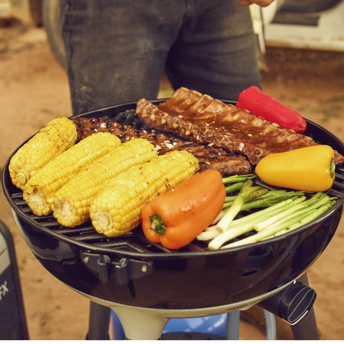 Cadac Carri Chef 40 - BBQ & Chef Pan Combo lifestyle image of full grill  with ribs, corn, peppers and spring onions . Someone's legs in black trousers can be seen behind. 