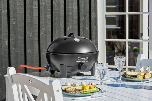 Cadac Citi Chef 40 Black - Urban & Camping Gas BBQ feature lifestyle image of BBq on table with plates of food in front of chairs and  wine glasses full with window in background