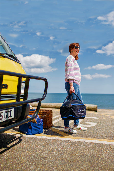 Cadac Citi Chef 40 Black - Urban & Camping Gas BBQ lifestyle image of woman in front of yellow van holding BBQ in carry bag with sea in background. 