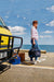 Cadac Citi Chef 40 Black - Urban & Camping Gas BBQ lifestyle image of woman in front of yellow van holding BBQ in carry bag with sea in background. 