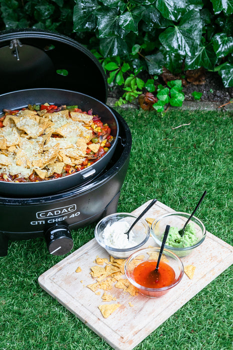 Cadac Citi Chef 40 Black - Urban & Camping Gas BBQ feature lifestyle image of BBQ on grass with nachos in and sauces on chopping board 
