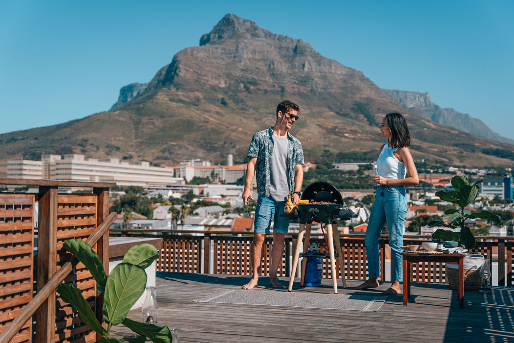 Cadac Citi Chef 40 FS Black - Urban & Camping Gas BBQ lifestyle image of BBq on deck with two people grilling in a city with mountains in background