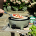 Cadac Pizza Lifter - BBQ Pizza Paddle lifestyle image of pizza being placed on citi chef 40 BBQ on outside table, with rolling pin to right side and Maldon salt next to cadac C500 cartridge. Pizza has sauce cheese and extra cherry tomatoes on. Out of focus paved garden with flower bushes in background. Hand on pizza lifter comes in the left side of picture. 
