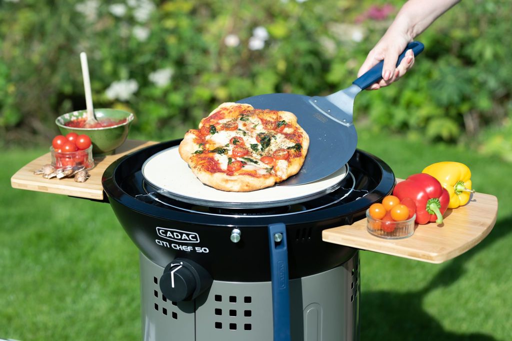 Cadac Pizza Lifter - BBQ Pizza Paddle lifestyle image of pizza cooked and being lifted off. The side of the Citi Chef 50 has red and yellow peppers and tomatoes one side and red tomatoes and a sauce in green bowl on the other wooded side table. Green grass and plants out of focus in the background