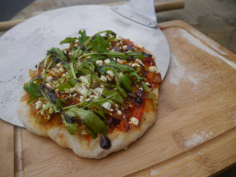 Cadac Pizza Lifter - BBQ Pizza Paddle lifestyle image, close up of the lifter with pizza on it with a cadac wooden chopping board below dusted with flour. Pizza has onions, balsamic, feta and rocket it. All on out of focus wooden table