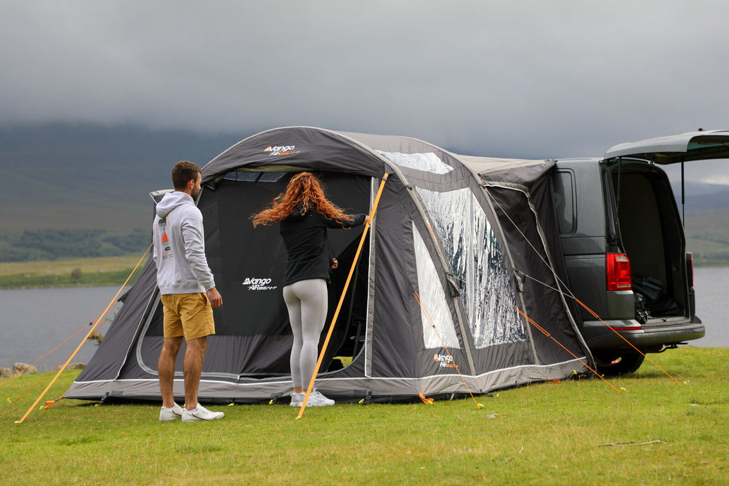 Vango Kela Pro Air Drive Away Awning - Low lifestyle image of awning pitched on VW camper  on grass with mountains and lake in background.  Fron t view with man and woman standing with backs to camera, woman zipping up front door. Trunk of van open. 