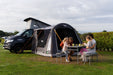 Vango Kela Pro Air Drive Away Awning - Low lifestyle image of awning pitched on VW camper  on campsite with green hedge in background.  front door unzipped and family of 4 on picnic bench out front