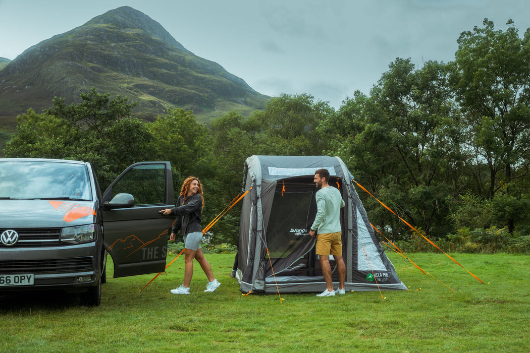 Vango Kela Pro Air Drive Away Awning - Low lifestyle image of awning pitched near VW camper on grass with mountains and tress in background. Side view of awning free standing with tunnel rolled up and man zipping up side door.  woman getting into van on left side. 