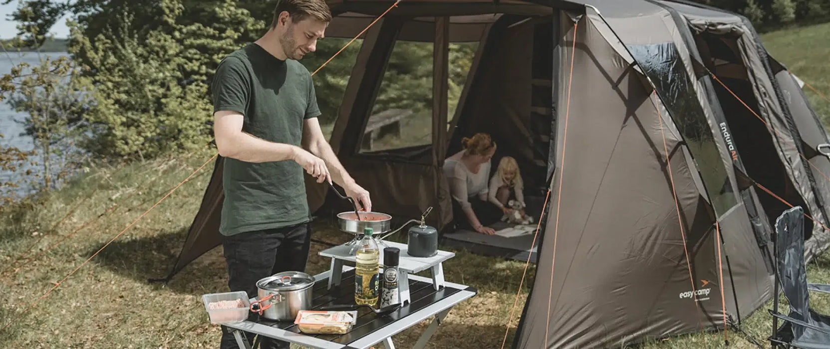 Easy Camp Moss Tour Stainless Steel Cook Set - lifestyle image, showing person using cook set with camping stove on table in front of tent with parent and child inside