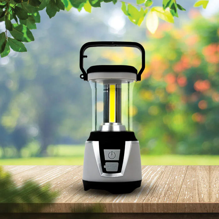 Leisurewize 3W COB LED Rechargeable Camping Lantern 1000 Lumens - product image, showing a portable LED camping lantern with a clear glass body, black handle, and silver and black base, shown lit on a wooden surface with a blurred garden background.