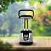 Leisurewize 3W COB LED Rechargeable Camping Lantern 1000 Lumens - product image, showing a portable LED camping lantern with a clear glass body, black handle, and silver and black base, shown lit on a wooden surface with a blurred garden background.