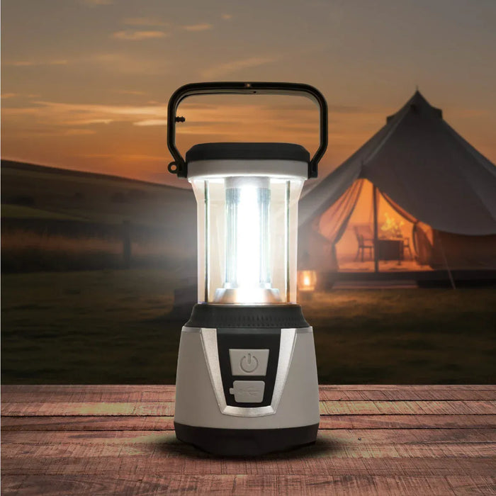 Leisurewize 3W COB LED Rechargeable Camping Lantern 1000 Lumens - product image, showing a portable camping lantern with a clear body, black handle, and illuminated light, placed on a wooden surface with a blurred campsite scene and tent in the background at sunset.