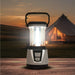 Leisurewize 3W COB LED Rechargeable Camping Lantern 1000 Lumens - product image, showing a portable camping lantern with a clear body, black handle, and illuminated light, placed on a wooden surface with a blurred campsite scene and tent in the background at sunset.
