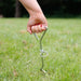 Leisurewize Dog Ground Anchor Screw Peg with 4m Tether Lead - lifestyle image, showing a hand with red nail polish holding a silver metal ground anchor stake with a ring attachment, set into a green grass lawn.