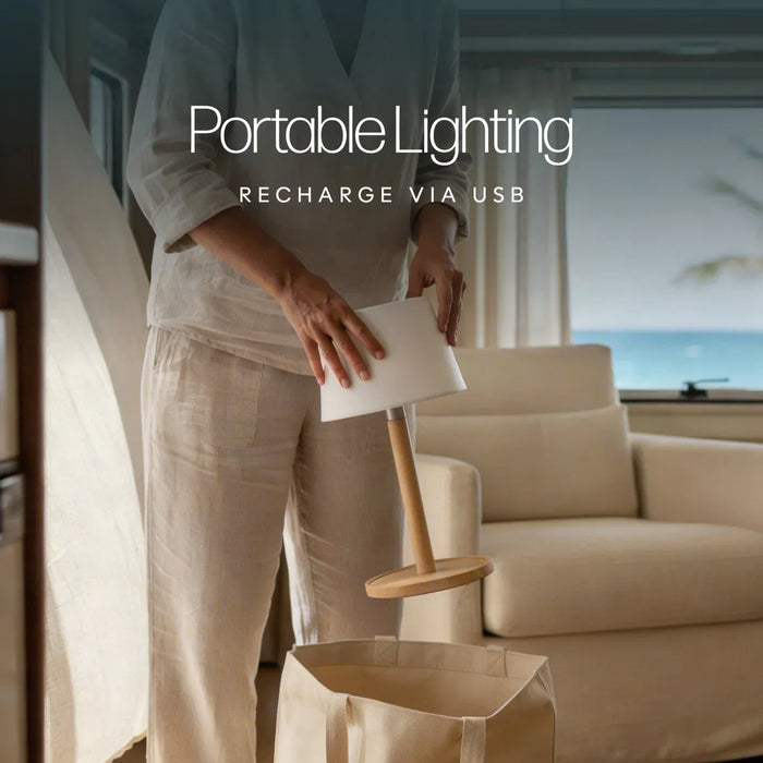 Leisurewize Orli Rechargeable Table Lamp – product image, showing a person in light beige clothing holding a portable table lamp with a white rectangular shade and a wooden base above a beige bag, with large text reading 'Portable Lighting' and 'RECHARGE VIA USB' over the scene. A sofa and window with ocean view are in the background.