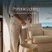 Leisurewize Orli Rechargeable Table Lamp – product image, showing a person in light beige clothing holding a portable table lamp with a white rectangular shade and a wooden base above a beige bag, with large text reading 'Portable Lighting' and 'RECHARGE VIA USB' over the scene. A sofa and window with ocean view are in the background.
