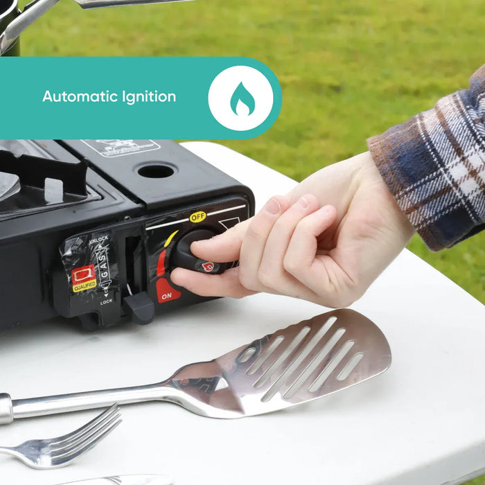 Leisurewize Single Burner Portable Gas Stove 2.5kW - product infographic, showing a close-up of a hand turning a dial on a black portable gas stove, with a teal banner reading 'Automatic Ignition' at the top. A metal spatula and fork rest on a white table outdoors, with grass visible in the background.