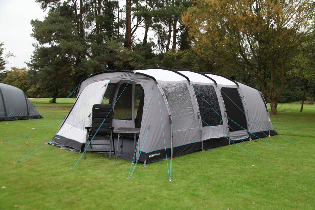 Outdoor Revolution Camp Star 600 DT- Poled 6-Berth Tunnel Tent Bundle lifestyle image of pitched on campsite with trees surrounding.. Side door has mesh zipped up and front door is half open of the right side with chair and table