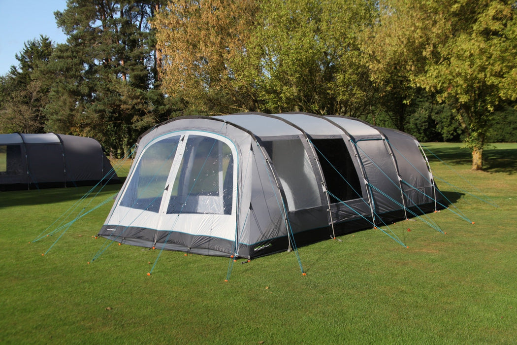 Outdoor Revolution Camp Star 600 DT- Poled 6-Berth Tunnel Tent Bundle lifestyle side view image of tent pitched on campsite with trees surrounding and another tent to the left. Image shows right side and front of the tent. The main front door is zipped up with all windows without curtains toggled up. Side door is closed