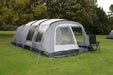 Outdoor Revolution Camp Star 600 DT- Poled 6-Berth Tunnel Tent Bundle lifestyle side view image of tent pitched on campsite with trees surrounding. The image shows left side of the tent which has no door on the side. the front door is half open on the right side with a table and chair sat on the groundsheet