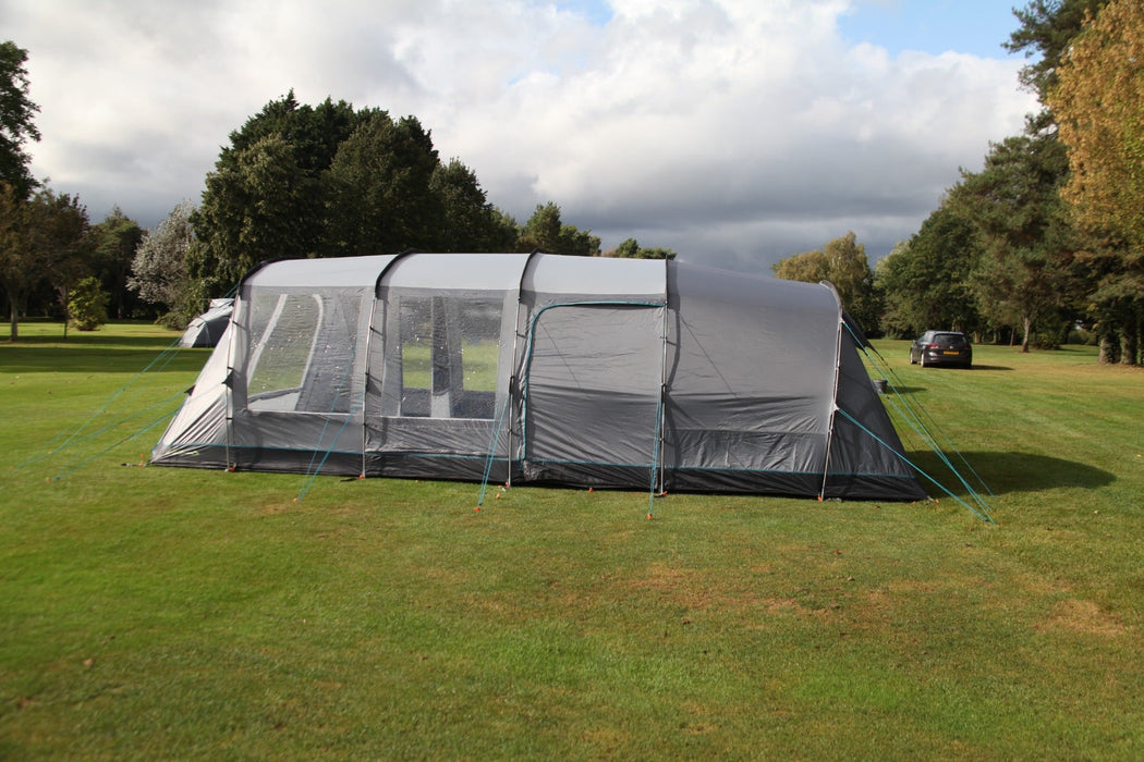 Outdoor Revolution Camp Star 600 DT- Poled 6-Berth Tunnel Tent Bundle lifestyle side view image of tent pitched on campsite with trees surrounding with car off to the right. the side door is closed and the front 2 window panels are open so you can see in and through the tent