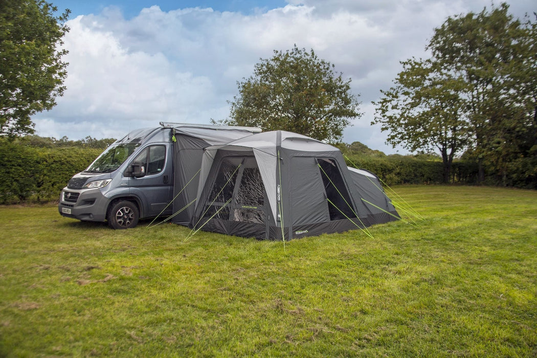 orda1623-04 A gray motorhome awning tent attached to the side of a gray van, pitched on a grassy lawn with trees and a cloudy blue sky in the background.