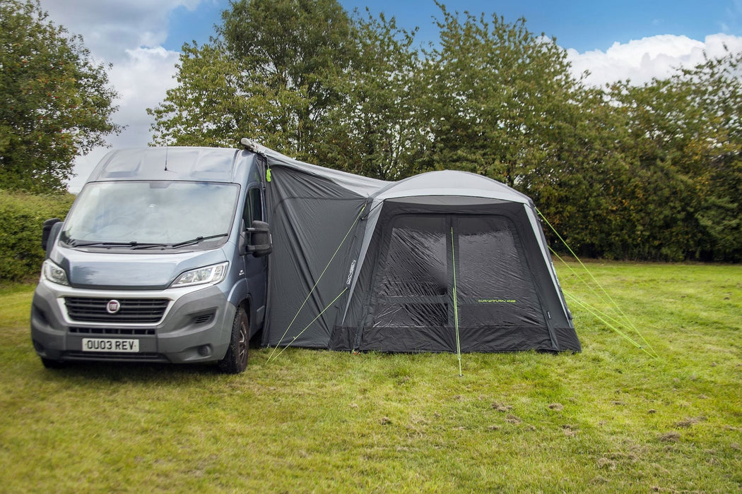 orda1623-08 A gray Camper World awning attached to the side of a silver Fiat van, pitched on a grassy field with trees and a blue sky in the background.