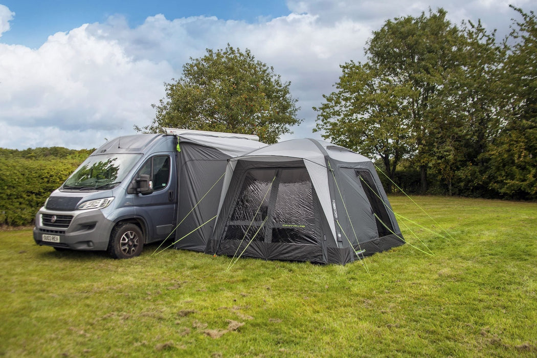 orda1623-09 A gray and black motorhome canopy tent attached to the side of a silver camper van, pitched on a grassy field with trees and a cloudy blue sky in the background.