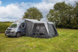 orda1623-09 A gray and black motorhome canopy tent attached to the side of a silver camper van, pitched on a grassy field with trees and a cloudy blue sky in the background.