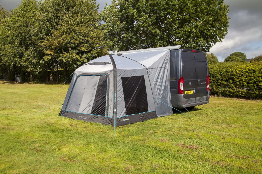 A gray and black pop-up caravan awning tent with mesh windows, shown attached to the side of a dark gray van on a grassy lawn with trees in the background.