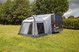 A gray and black pop-up caravan awning tent with mesh windows, shown attached to the side of a dark gray van on a grassy lawn with trees in the background.