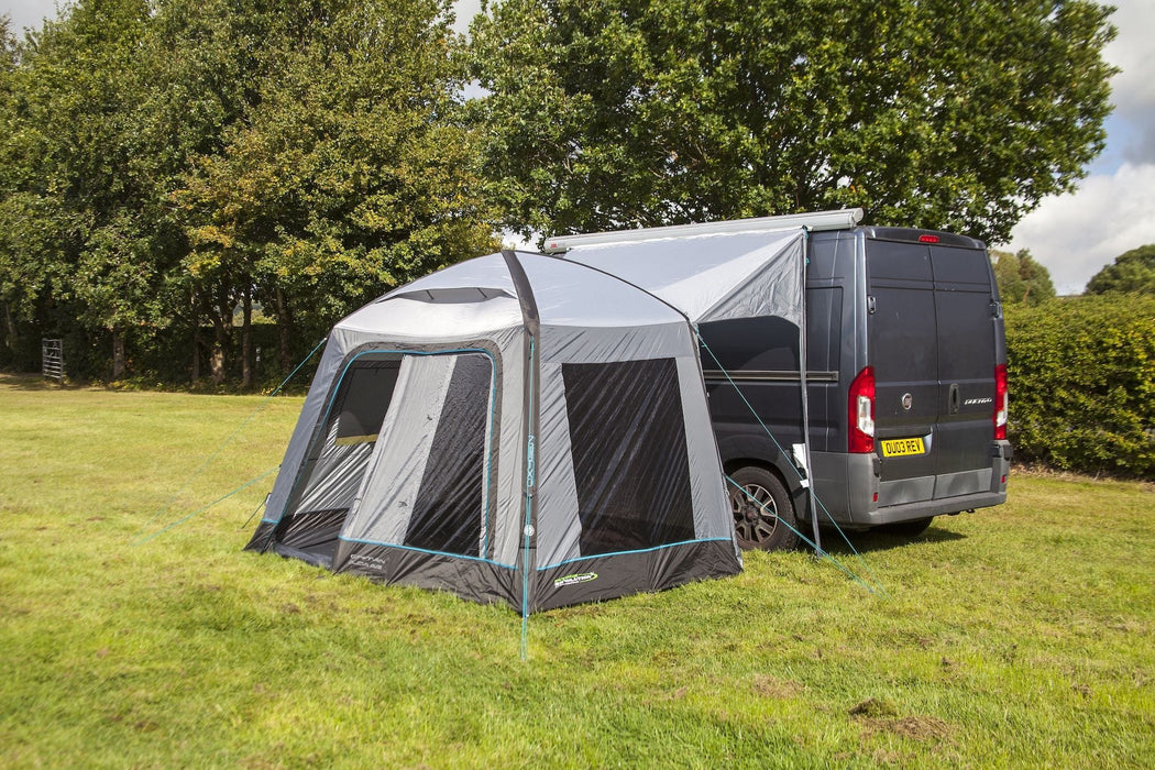 Van with a gray and black awning extended outdoors on a grassy area with trees in the background.