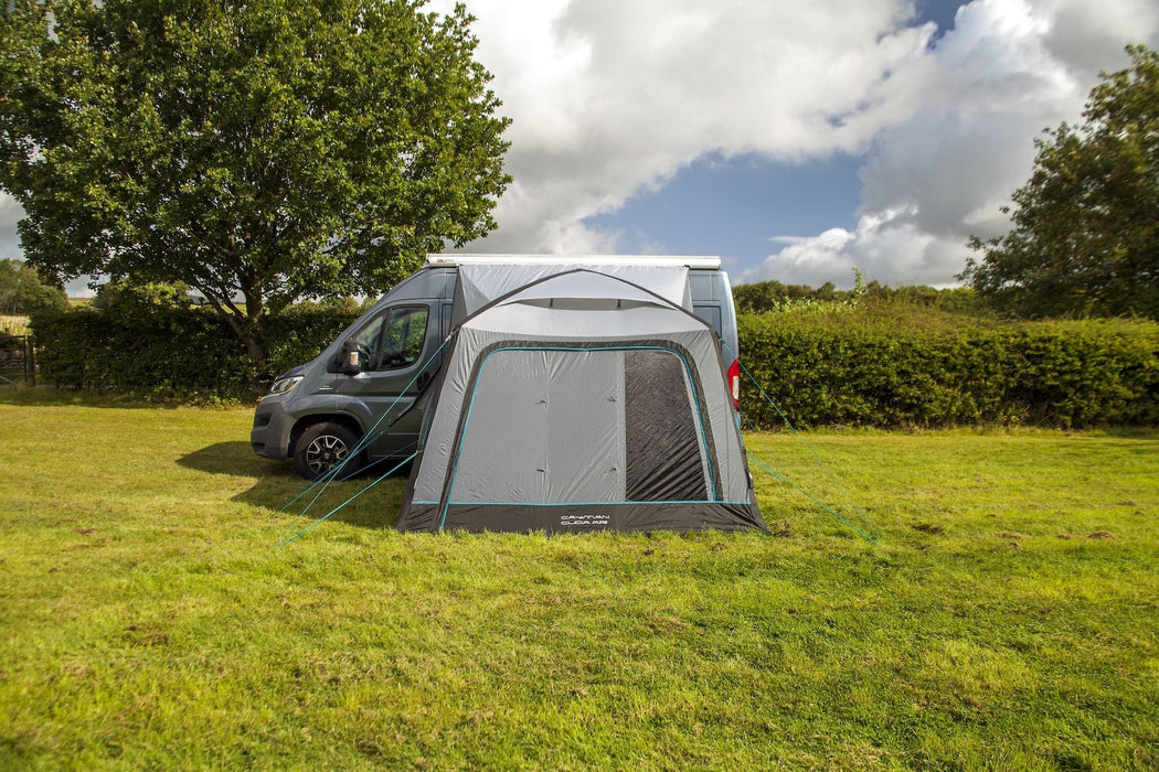 Van with an attached tent on a grassy field under a blue sky with clouds.