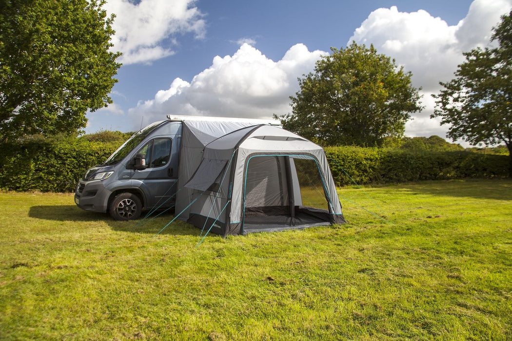 A gray and white motorhome awning tent pitched over the side of a dark gray van on a grassy lawn, with trees and a blue sky with clouds in the background.