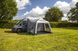 A gray and white motorhome awning tent pitched over the side of a dark gray van on a grassy lawn, with trees and a blue sky with clouds in the background.