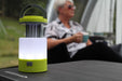 Outdoor Revolution Lumi-Mosi Recharageable Lantern & Mosquito Killer - lifestyle image, showing lit lantern, with mosi-killer compartment extended, sat on table outside with person sat in background