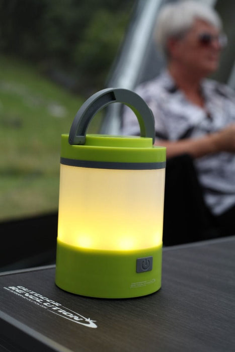 Outdoor Revolution Lumi-Mosi Recharageable Lantern & Mosquito Killer - lifestyle image, showing lit lantern, without mosi-killer compartment extended, sat on table outside with person sat in background
