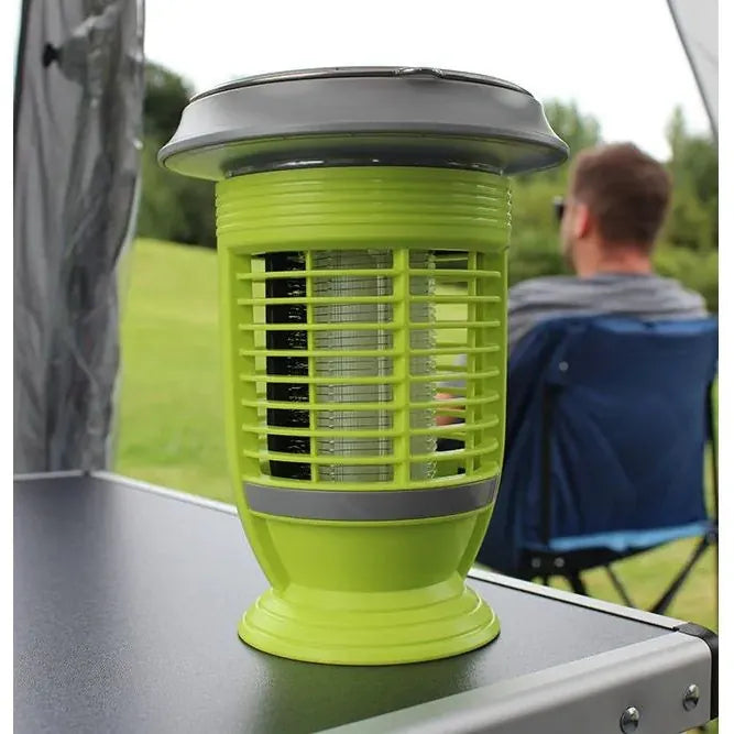 Outdoor Revolution Lumi-Solar Mosi Killer Lantern - product lifestyle image, showing a bright lime green and gray electric bug zapper lamp sitting on a dark camping table, with a blurred man sitting in a blue folding chair in the background outdoors.