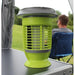 Outdoor Revolution Lumi-Solar Mosi Killer Lantern - product lifestyle image, showing a bright lime green and gray electric bug zapper lamp sitting on a dark camping table, with a blurred man sitting in a blue folding chair in the background outdoors.