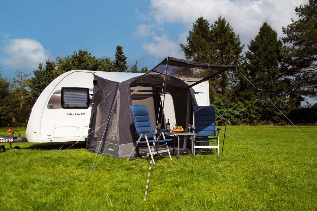 Outdoor Revolution Porch Pro 260 Inflatable Caravan Awning - product image, showing awning pitched and attached to caravan in a field surrounded by trees, with front door raised open as a canopy, with chairs and table in front of awning