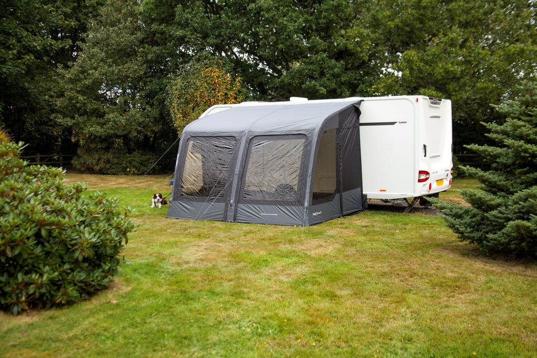 Outdoor Revolution Sport Lite Air 330 Inflatable Caravan Awning - product image, showing distant front-right side view of awning attached to caravan pitched in field with surrounding trees and bushes, with dog lying on grass to left-hand side of awning