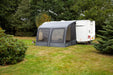 Outdoor Revolution Sport Lite Air 330 Inflatable Caravan Awning - product image, showing distant front-right side view of awning attached to caravan pitched in field with surrounding trees and bushes, with dog lying on grass to left-hand side of awning