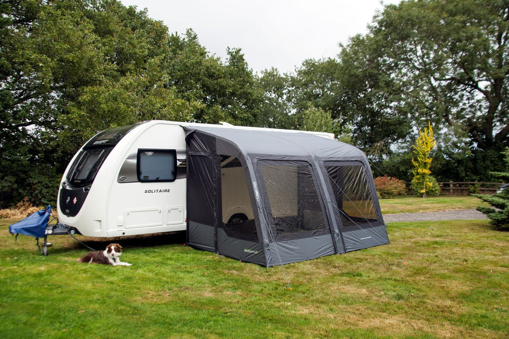 Outdoor Revolution Sport Lite Air 330 Inflatable Caravan Awning - product image, showing front-left side view of awning attached to caravan pitched in field with surrounding trees, and dog lying on grass to left-hand side of awning