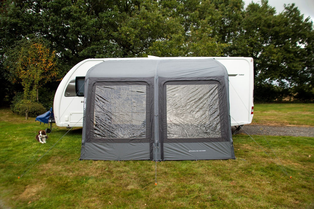 Outdoor Revolution Sport Lite Air 330 Inflatable Caravan Awning - product image, showing front-on view of awning attached to caravan pitched in field with surrounding trees, and dog lying on grass to left-hand side of awning